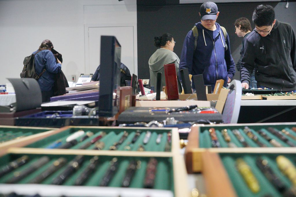 A collection of pens rest in green trays, out of focus in the foreground. In the background, visitors look down at items on the exhibitor's other tables.