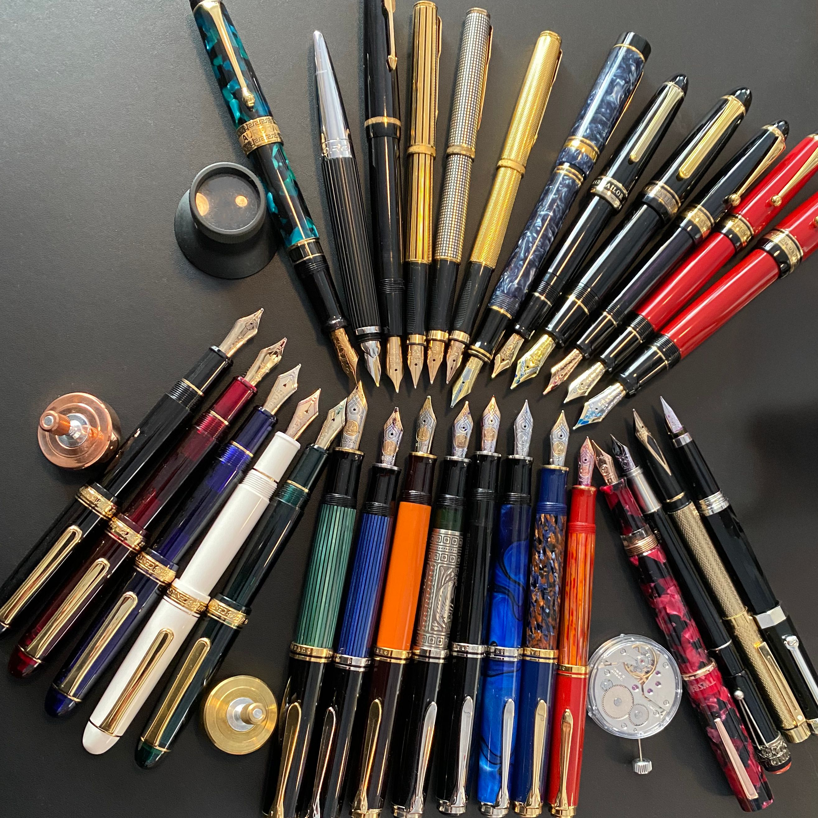 Thirty fountain pens scattered on a black leather desk mat with various other writing paraphernalia