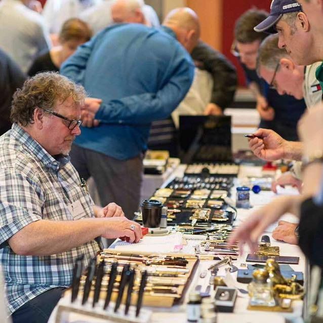 a man sits at a table covered in pens for sale, helping curious customers