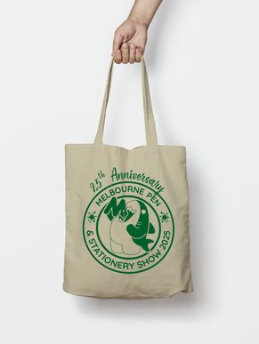 A hand holds a cream-coloured canvas tote bag with a green logo on it: a cartoon shark gobbles up the Melbourne Pen & Stationery Show logo inside a circular band with ink splotches and the text: "Melbourne Pen & Stationery Show 2025". At the top is the text "25th anniversary" in a handwritten script.