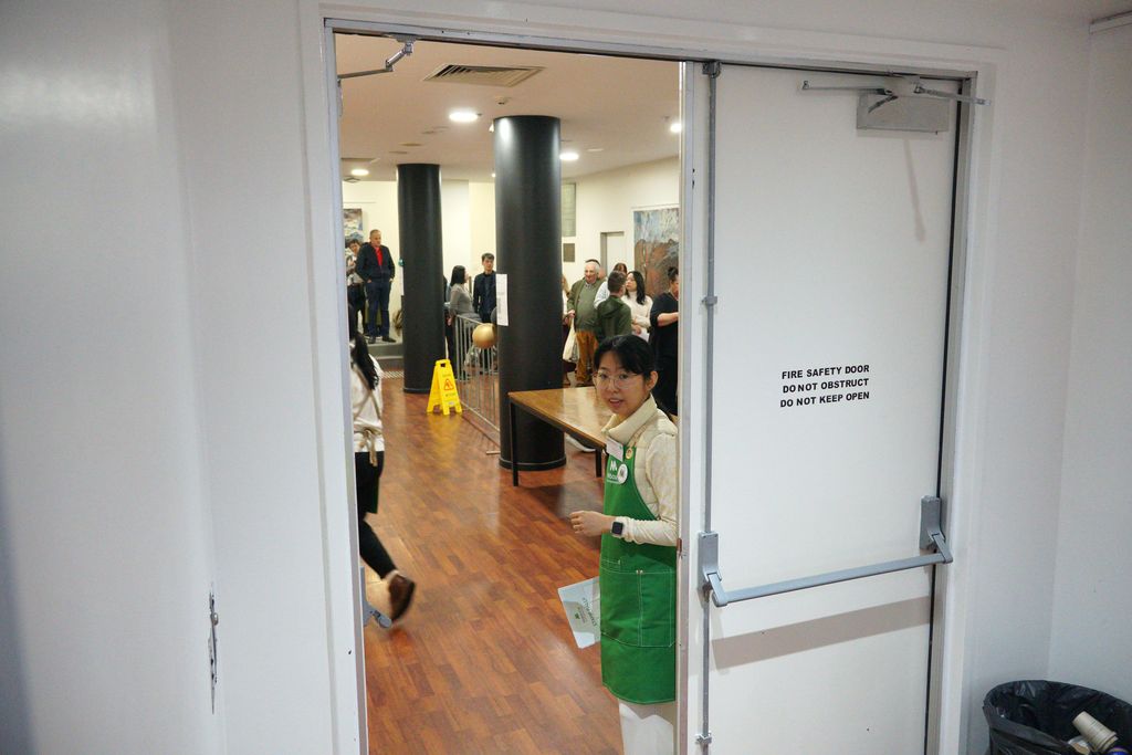 Looking out the hall doorway into the foyer at a volunteer in a green apron and the line of queued guests waiting to enter