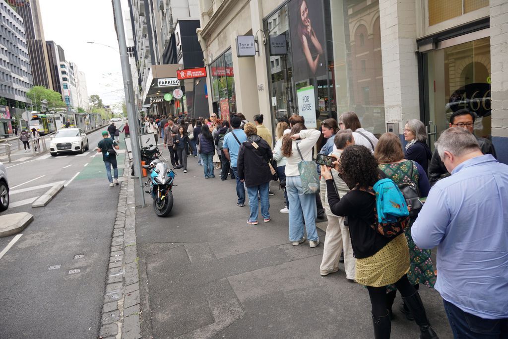 A long queue of people down the sidewalk. One steps out to take a photo with their phone.