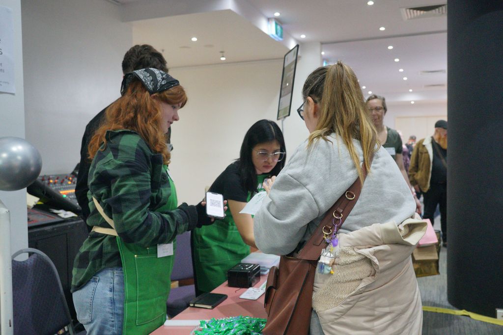 A volunteer wearing a green apron shows a phone with a QR code to a visitor