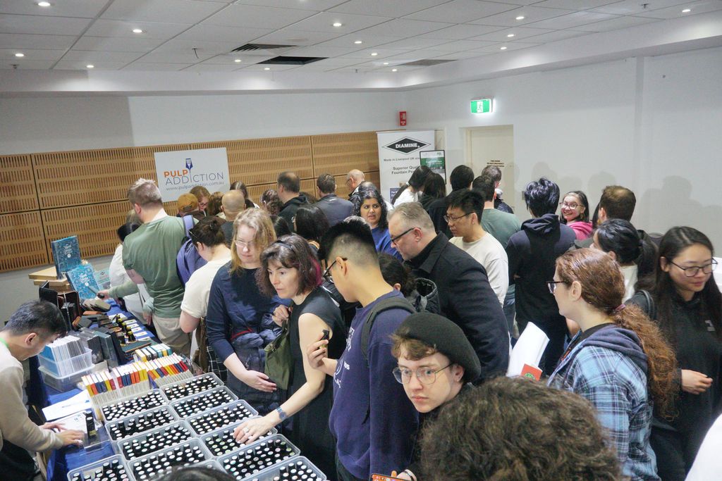 A crowd of people in the back corner of the show floor, with a table full of Robert Oster ink bottles in the foreground