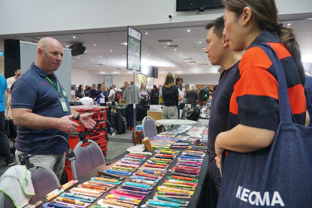 An exhibitor helps two visitors over a table full of colourful resin pens